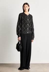Black knitted cardigan with a diamond pattern, silver threading, and buttoned front. Paired with loose black trousers and a small black handbag.