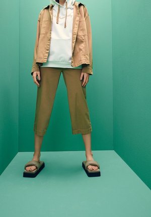 Persoon draagt beige cropped broek, witte hoodie, beige jas en sandalen met plateauzool, staand tegen een teal gekleurde muur en vloer.