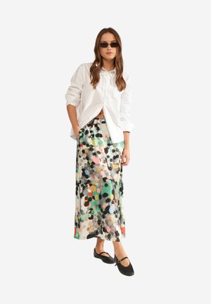 Kvinde iført sorte solbriller, hvid skjorte med knapper, farverig polkaprikket midinakskørt og sorte Mary Jane-flade sko, stående med hænderne i lommerne.