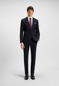 Marineblauwe blazer met geknipte revers, gecombineerd met slim-fit broek. Lichtroze overhemd eronder, bordeauxrode stropdas en een gestreepte pochet. Zwarte schoenen.