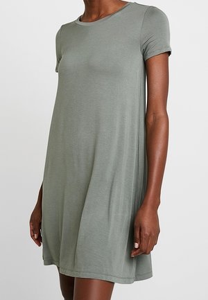 Woman wearing a short-sleeve, knee-length, loose-fitting light gray-green dress against a plain background.