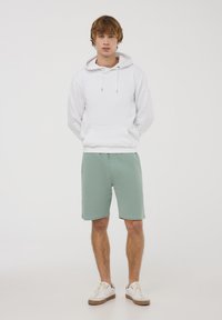 Felpa bianca in cotone con tasca frontale, abbinata a pantaloni corti verdi chiari. Il modello indossa sneakers bianche con dettagli color sabbia. Design semplice e casual.