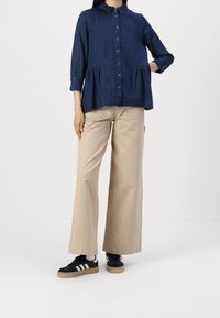 Denim button-up shirt with a collar, gathered waist, and three-quarter sleeves. Paired with wide beige trousers and black sneakers.