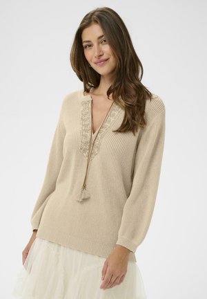 Jeune femme aux longs cheveux bruns portant un pull en tricot beige avec broderies et détails à pompons, associé à une jupe en tulle blanche.
