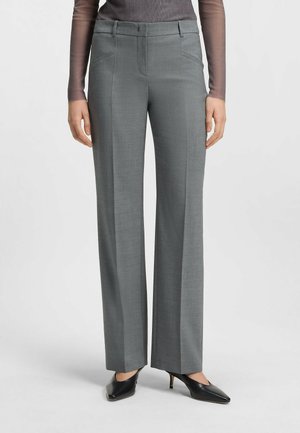 Kobieta ubrana w szare spodnie z prostymi nogawkami z przednimi kieszeniami oraz czarne szpilki z wąskim noskiem, stojąca na jednolitym tle.