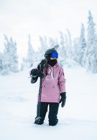 Lyserød sweatshirt med logo, sorte skibukser, sort hjelm og blå skibriller, holder et snowboard. Sneklædt landskab i baggrunden.