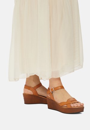 Sandalias de cuña de cuero marrón con tiras entrelazadas y una hebilla ajustable en el tobillo. El tacón de cuña presenta un acabado texturizado. Combinadas con un vestido ligero.