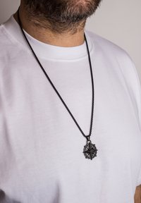 Pendentif de compas en métal noir sur une fine chaîne, porté sur un t-shirt blanc. Le pendentif présente des détails complexes et un design en étoile au centre.