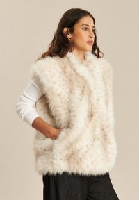 Nepbont vest in crème met een lichte textuur, voorzien van een v-hals en zijzakken, gedragen over een lange mouwen witte top.