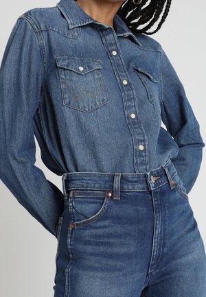 Personne portant une chemise en jean bleu ajustée avec des poches boutonnées et un jean bleu taille haute, debout les mains derrière le dos.