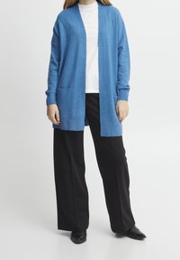 Jasnoniebieski kardigan z miękkiego materiału, z otwartym przodem, długimi rękawami i dwoma przednimi kieszeniami, w połączeniu z czarnymi spodniami o szerokich nogawkach.