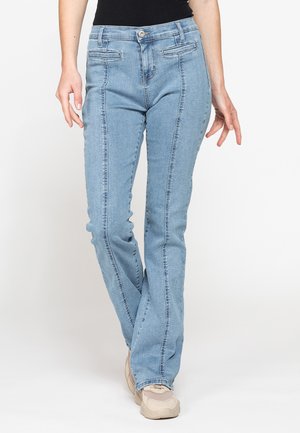 Donna che indossa jeans azzurri a gamba dritta e sneakers beige, in piedi davanti a uno sfondo semplice.