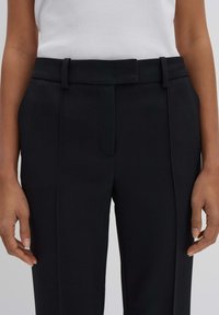 Pantaloni neri su misura con una texture liscia, dotati di una cintura, pieghe e una vita aderente. Il tessuto appare strutturato e formale.