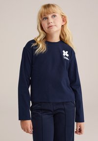 Jūros mėlynos ilgos rankovės marškiniai iš raukšlėto audinio, su balta logo krūtinėje. Pora su tamsiai mėlynomis kelnių.
