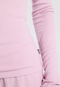 Pink long-sleeve top in ribbed fabric with fitted design. Features a subtle logo tag on the side and a smooth texture.