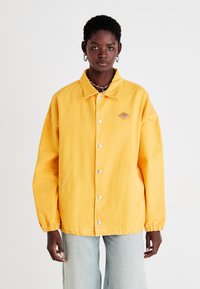 Gul jakke i avslappet passform, med knappelukking, to sidelommer, elastiske cuffer og et lite logo-patch på brystet.