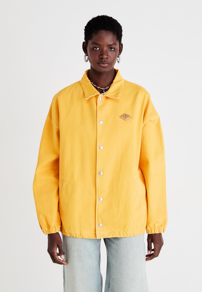Gul jakke i avslappet passform, med knappelukking, to sidelommer, elastiske cuffer og et lite logo-patch på brystet.