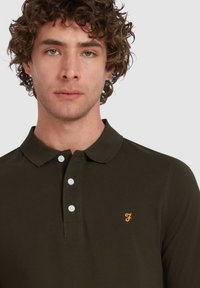 Donkergroene polo shirt van katoen, met een tweeknoopsluiting, witte knopen en een klein oranje logo op de linkerborst.