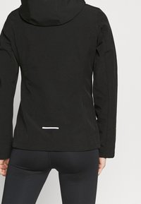 Chaqueta impermeable negra con capucha, que presenta una textura suave y un acento reflectante blanco cerca del dobladillo. Diseñada para la funcionalidad y la comodidad.