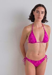 Ružičasti bikini set s trokutastim vrhom i gaćicama koje se vezuju sa strane, s teksturiranim, uzorkovanim dizajnom. Tkanina izgleda glatko i lagano.