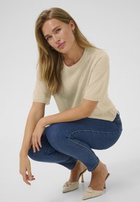 Haut en tricot beige à manches courtes, col rond. Associé à un jean skinny bleu foncé et des mules à talons beiges avec un détail en nœud.