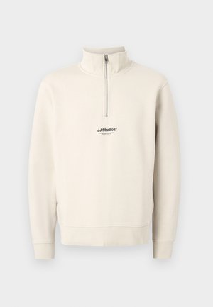 Beige quarter-zip sweatshirt with long sleeves and ribbed cuffs. Features a small black "JJ Studios" logo on the chest. Made of soft material.