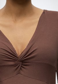 Κοντινό πλάνο ενός ατόμου που φοράει καφέ ριμπ πουκάμισο με στριμμένο κόμπο στο βαθύ V-λαιμό.