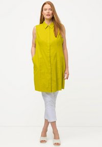 Túnica sin mangas de color verde lima con cuello, botones en la parte delantera y bolsillos laterales. Combinada con pantalones blancos de corte cropped y zapatos de tacón cuadrado.