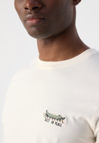Tricou din bumbac de culoare bej deschis, cu un mic crocor prăjit brodat și cu textul "ODIHNEASCĂ ÎN PACE." Guler rotund simplu și mâneci scurte.