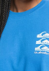 Niebieska bawełniana koszulka z białym nadrukiem przedstawiającym fale i góry. Okrągły dekolt z widocznym przeszyciem.