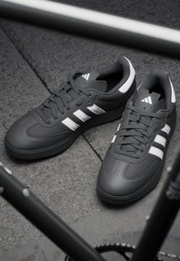 Szare buty sportowe z białymi paskami, wykonane z gładkiego materiału syntetycznego, z okrągłym noskiem, sznurowanymi zapięciami i teksturowaną czarną podeszwą.