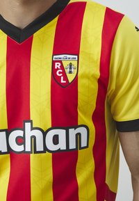 Rød og gul stribet fodboldtrøje med en sort V-hals og ærmeaccentueringer. Har et Racing Club de Lens-logo og et stort hvidt sponsorlogo.
