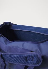 Tamno plavi torba za teret s patentnim zatvaračem na vrhu, podstavljenim mrežastim rukohvatom i unutarnjim pregradama; ima izdržljiv, teksturiran završni materijal.