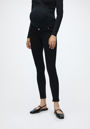 Femme enceinte portant un jean de maternité noir avec un panneau élastique au niveau du ventre et des chaussures plates noires sur un fond uni.