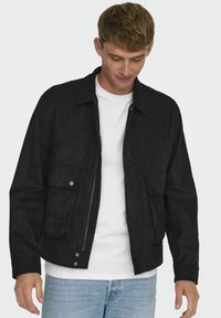 Svart mockajacka med dragkedja fram, två framfickor och krage, bärs över en vit t-shirt, tillsammans med ljusa denimjeans.