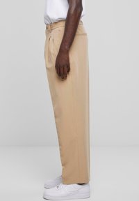 Beige broek met een rechte snit, plooien aan de voorkant en een zachte stof. Gecombineerd met witte sneakers, tegen een neutrale achtergrond.