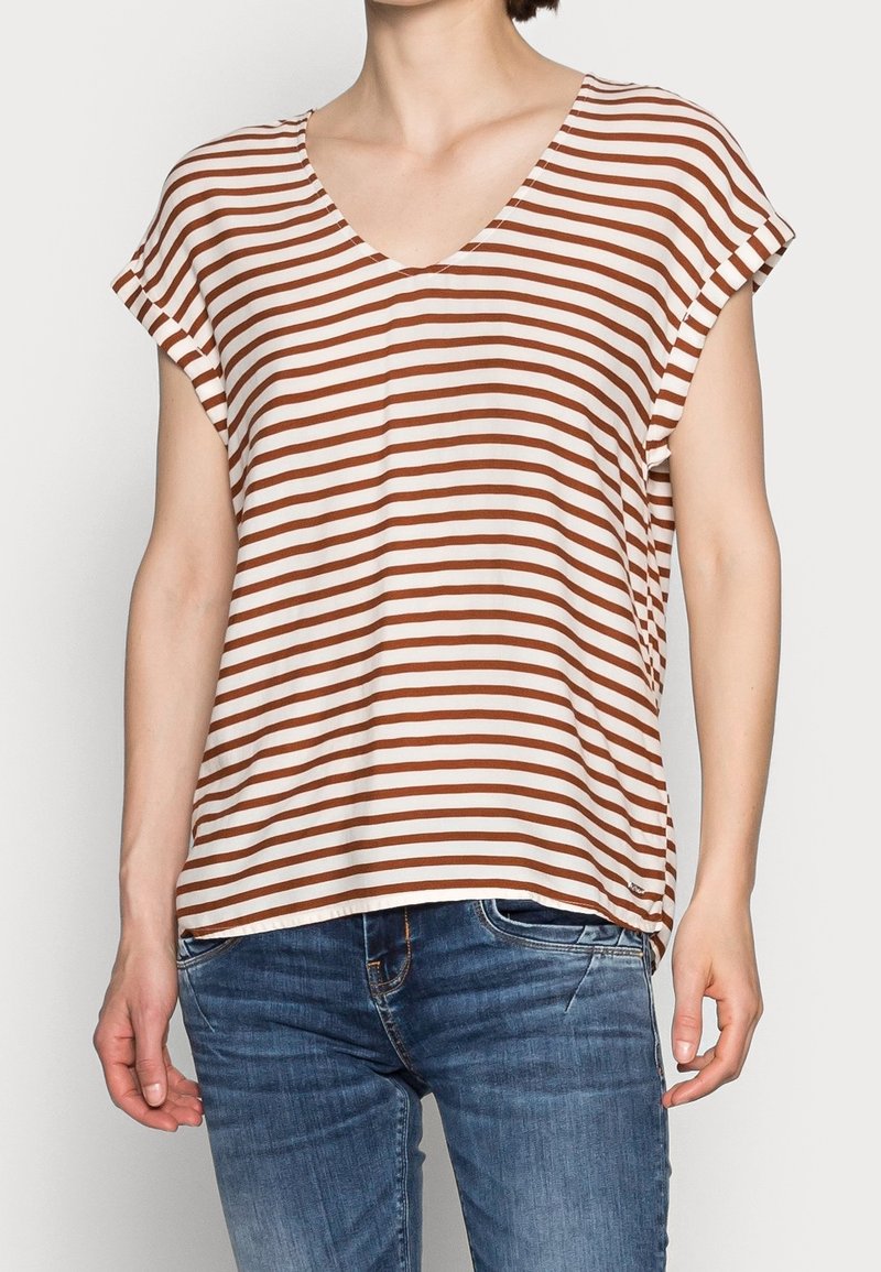 Mujer vestida con una blusa de cuello en V de manga corta a rayas marrón y blanca, combinada con jeans de mezclilla azul, de pie contra un fondo liso.