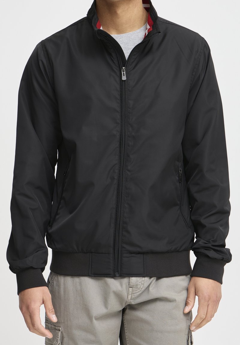 Mand iført en sort zip-jakke med ribbede manchetter og nederkant over en grå skjorte, parret med lysegrå cargo-bukser.