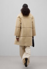 Beige shearling-kappa med fyrkantigt panelmönster, vida ärmar och en synlig söm. Kombinerad med krämfärgade byxor och håller en svart väska.