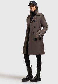 Cappotto marrone con design doppiopetto, bottoni neri e cintura. Abbinato a leggings neri e stivali neri spessi. Un cappello completa il look.