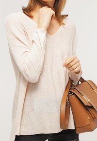 Jasnobeżowy sweter na długi rękaw wykonany z miękkiego, teksturowanego dziania, z dekoltem w serek i przednią kieszenią, w zestawieniu z brązową torebką na ramię.