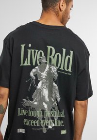 Black cotton t-shirt featuring a large graphic of a cowboy on horseback with text “Live Bold” and “Live tough, push hard, exceed every line.”