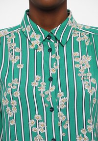 Green shirt featuring vertical navy stripes and white daisies; includes a collar and buttons along the front.