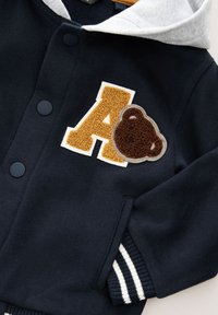 Felpa blu navy con cappuccio grigio, caratterizzata da una "A" gialla testurizzata e una toppa a forma di orso marrone, rifinita con polsini a righe bianche.