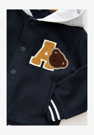 Tengerészkék pulóver szürke kapucnival, egy texturált sárga "A" betűvel és egy barna medve folttal díszítve, fehér csíkos mandzsettákkal befejezve.