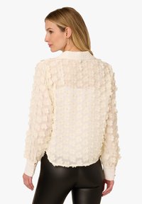 Cream-colored blouse with floral texture, sheer fabric, long sleeves, and a collared neckline. Features a solid back and a relaxed fit.