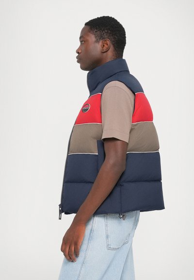 Gewatteerde vest in marineblauw, rood en kaki kleuren; heeft een hoge kraag, ritssluiting en logo patch op de borst. Getextureerde stof met horizontale lijnen.