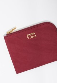 Czerwona portmonetka z złotym suwakem i logo "BIMBA Y LOLA." Ma gładką teksturę i prostokątny kształt.