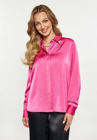 Blusa rosa satinata con scollo a collare, bottoni frontali, maniche lunghe e spilla decorativa sul colletto. Tessuto morbido e vestibilità rilassata.