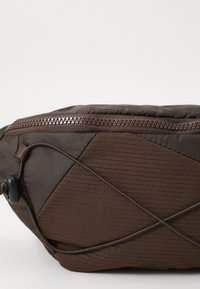 Brown nylon bag with a textured surface, zip closure, and mesh accents. Features a curved shape and bungee cord detail.